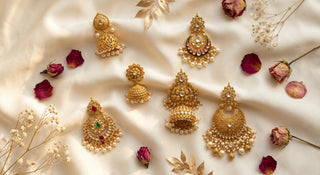 A top-down flat lay view of six assorted gold-plated jhumka earrings for women, scattered organically across soft ivory silk fabric and decorated with dried red rose petals