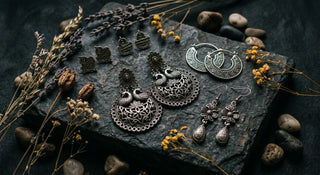 Moody flat lay of tribal-style oxidised silver earrings featuring peacock motifs and geometric patterns on a dark stone surface with dried wildflowers