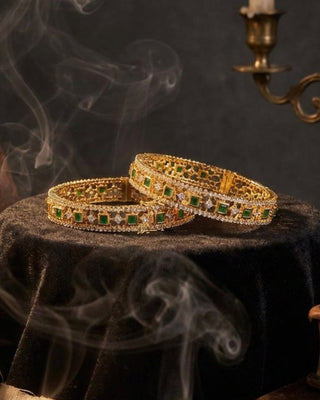 Gold bangles with green gemstones on a dark surface with smoke and candles in the background