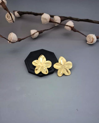 Gold flower-shaped earrings on a black stand with decorative branches in the background.