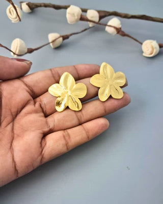 Gold flower-shaped earrings held between fingers with a light blue background