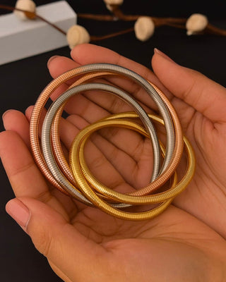 Hand holding three metallic bangles in gold, silver, and bronze on a dark background.