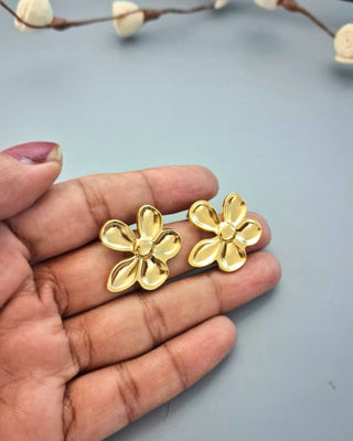 Gold flower-shaped earrings held in a hand against a light blue background.